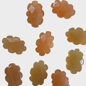 May include: A collection of nine, light orange, flower-shaped gemstones with faceted surfaces. The gemstones have a translucent quality and varying shades of orange and yellow. The gemstones are scattered on a white background.