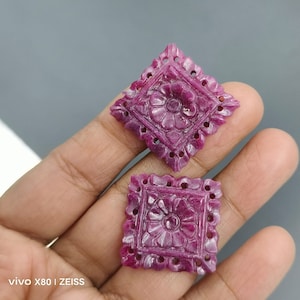 Puede incluir: Dos cuadrados de piedras preciosas rosadas talladas con intrincados diseños florales. Las piedras preciosas están engastadas en una forma cuadrada con un diseño de flor en el centro. Las flores tienen un centro elevado y pétalos tallados con detalle.