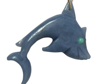 May include: A blue shark pendant carved from stone, with a turquoise eye and a gold-colored chain. The shark is stylized with fins and a tail, and is suspended from a thin, gold-colored chain.