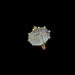 May include: A clear, carved crystal ring in the shape of a scarab beetle. The ring has a gold-colored band and small gold accents around the beetle's edges. The beetle is approximately 1 inch long.