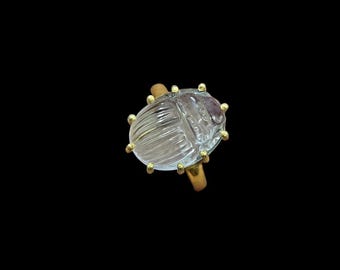 May include: A clear, carved crystal ring shaped like a scarab beetle. The ring features a gold-coloured band and small gold accents. The beetle is approximately 2.5 cm long.