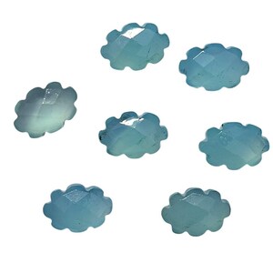 May include: Seven light blue, cloud-shaped gemstones with faceted surfaces are scattered against a white background. The gemstones have a translucent quality, and their edges are softly rounded. The overall effect is delicate and ethereal.