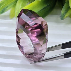 May include: A faceted, translucent purple ring with a geometric, multi-sided cut. The ring is held by a pair of metal tweezers, showcasing its transparent, deep violet hue and sharp, angular edges that reflect light across its polished surface.