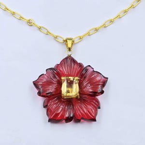 May include: A gold chain necklace featuring a carved deep red flower pendant. The center of the flower is set with a rectangular yellow gemstone held by gold prongs. The petals have detailed carved lines radiating from the center stone.