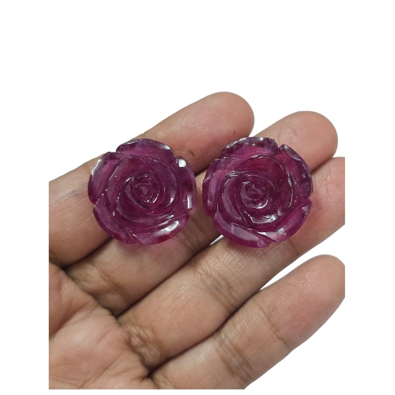 May include: Two carved ruby rose gemstones. The gemstones are a deep, rich red color and are intricately carved to resemble blooming roses. The details of the petals are clearly visible, showcasing the craftsmanship. The gemstones are set against a white background.