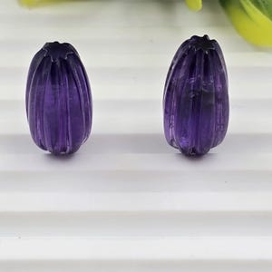 May include: Two carved amethyst gemstone beads. Each bead is a deep purple color, with vertical grooves and a flower-like top. The beads are oval-shaped and appear to be polished.
