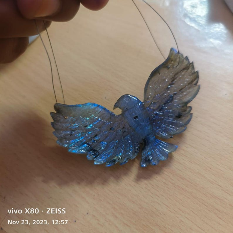 Puede incluir: Colgante de labradorita tallada con forma de p&aacute;jaro con alas extendidas. El p&aacute;jaro presenta un dise&ntilde;o detallado con tonos iridiscentes azules y verdes. El colgante est&aacute; suspendido por finos hilos, listo para ser usado como joya.