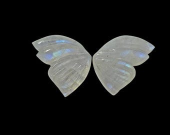 May include: Two carved, translucent moonstone butterfly wings against a black background. The wings are detailed with carved veins and display iridescent flashes of blue and green. The wings are symmetrical and appear to be a pair.