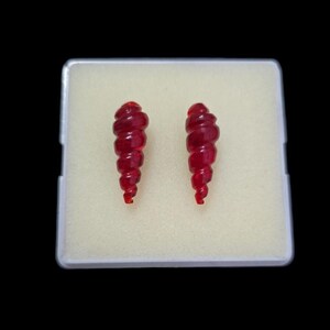 May include: Two red, spiral-shaped earrings are displayed in a white, rectangular case. The earrings have a textured, ribbed design and a glossy finish. The background is black.