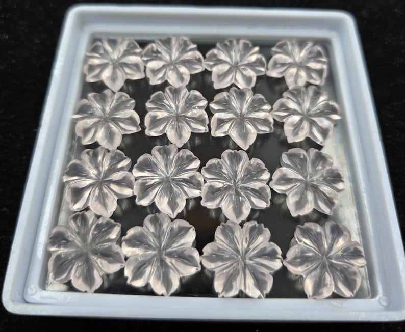 K&ouml;nnte beinhalten: Eine Sammlung von 15 klaren Quarz-Blumencabochons in einer wei&szlig;en Plastik-Schale. Die Cabochons sind durchscheinend und haben einen leicht rosafarbenen Farbton.