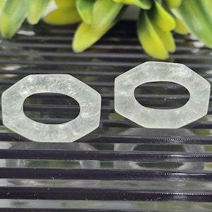 May include: Two light green, hexagonal gemstone pendants with oval cutouts. The pendants are translucent and appear to be made of a crystalline material. The background features a dark, ridged surface with blurred green and yellow foliage.