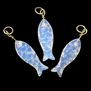 May include: Three opalescent fish pendants with gold-colored loops and small pink accents. The fish have a scale pattern and are set against a black background. These pendants are likely jewelry.