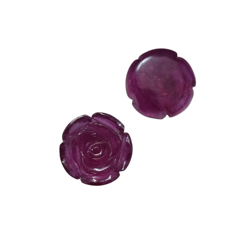 May include: Two carved rose-shaped gemstones in a deep magenta hue. One rose is fully detailed with petals, while the other is a simpler, rounded design. The gemstones are set against a plain white background.
