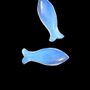 May include: Two light blue, fish-shaped decorative pieces. The fish have a translucent appearance and are set against a black background. The fish have a rounded body and a small tail fin.