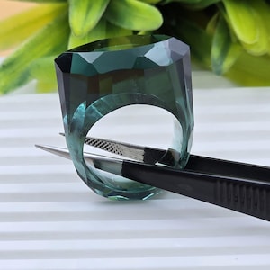 May include: A translucent, dark teal ring with a faceted, geometric outer surface. The ring is held by a pair of black metal tweezers, showcasing its thick, solid construction and deep green-blue color profile with clear, sharp edges.