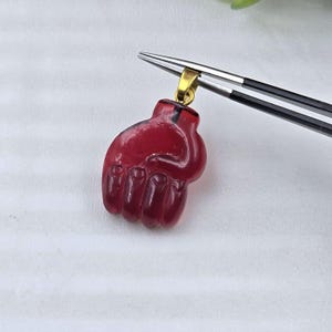 Puede incluir: Un colgante de piedra preciosa translúcida de color rojo intenso tallado con forma de puño cerrado. El colgante tiene una anilla de metal dorado en la parte superior, sostenido por unas pinzas de metal plateado. El tallado muestra dedos.