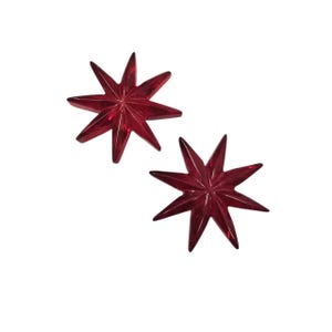 May include: Two star-shaped decorative objects in a deep red hue. Each star has eight pointed tips and a faceted surface, creating a sparkling effect. The objects are set against a plain white background.