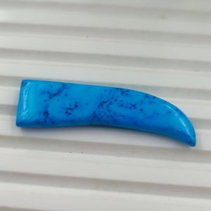 May include: A curved, tooth-shaped cabochon with a bright turquoise blue colour and dark, irregular veining throughout the surface. The object has a smooth, polished finish and a flat base, tapering to a point at one end with a wide, flat edge.