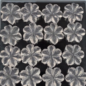 K&ouml;nnte beinhalten: Eine Sammlung von 15 klaren Quarz-Blumen-Cabochons, jede mit einem F&uuml;nf-Bl&uuml;ten-Design, angeordnet in einem Gittermuster in einer grauen Schale.