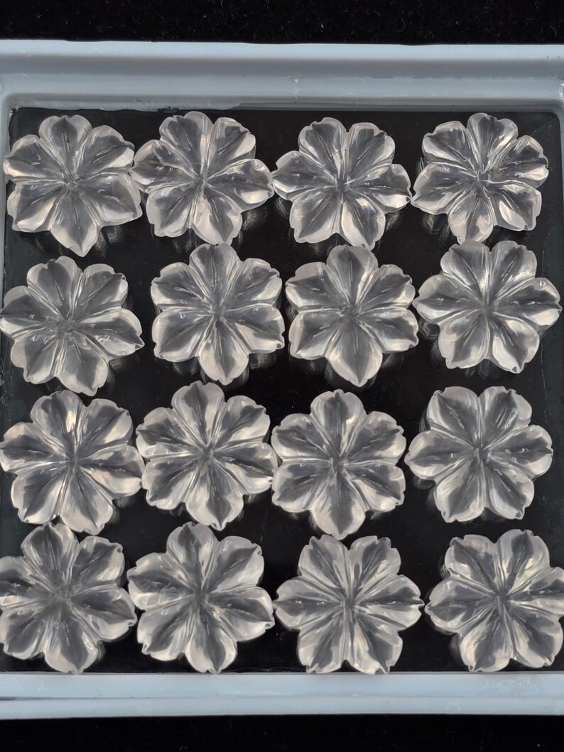 K&ouml;nnte beinhalten: Eine Sammlung von 15 klaren Quarz-Blumen-Cabochons, jede mit einem F&uuml;nf-Bl&uuml;ten-Design, angeordnet in einem Gittermuster in einer grauen Schale.