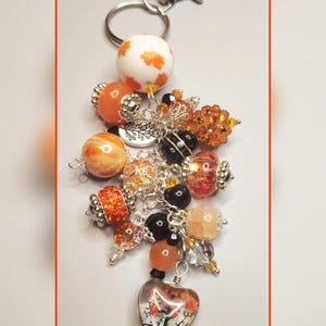 May include: A decorative keychain with a silver clasp and ring. The keychain features a variety of orange, white, and black beads, including a heart-shaped charm with a tree design. The beads are accented with silver-colored metal.