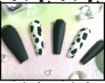 May include: A set of matte black and cow print press-on nails. Some nails have a gold glitter accent at the cuticle. The nails are long and coffin-shaped, with a glossy finish. The cow print nails have a white base with black spots.