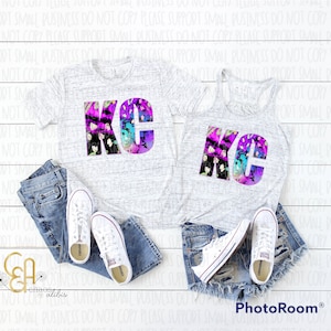 Könnte beinhalten: Zwei weiße Shirts mit einem Grafikdruck der Buchstaben "KC" in einem farbenfrohen, abstrakten Design. Die Shirts liegen auf einer weißen Holzoberfläche mit einem Paar weißen Sneakers und einer blauen Jeans-Shorts.