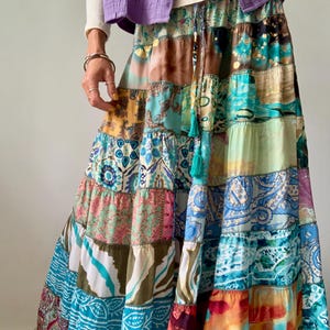 Könnte beinhalten: Maxirock im Patchwork-Design mit verschiedenen Stoffen in Türkis, Braun, Rosa und Blau. Der Rock hat einen Stufen-Look und einen Kordelzug in der Taille. Dazu ein lila Oberteil und ein weißes Langarmshirt.