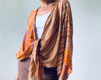 Pode incluir: Top estilo quimono listrado laranja e castanho com fecho de laço. O top apresenta um design listrado e detalhes florais, combinado com um top branco e uma minissaia castanha. Pulseiras e anéis prateados completam o look.