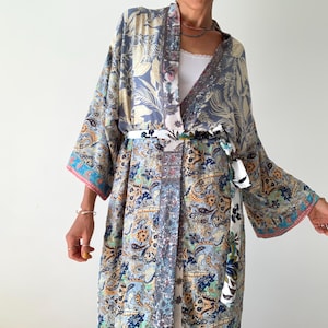 May include: A long, flowing kimono robe with a paisley and floral pattern in shades of blue, gold, and teal. The robe has wide sleeves and a matching belt. The edges are trimmed with pink and blue accents. The robe is worn open, revealing a white top.