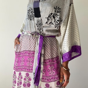 May include: A patterned kimono-style robe in white, black, and purple. The robe has a mix of floral and swirl patterns, with a purple belt to cinch the waist. The sleeves and hem are trimmed with a solid purple border. The robe is made of a lightweight fabric.