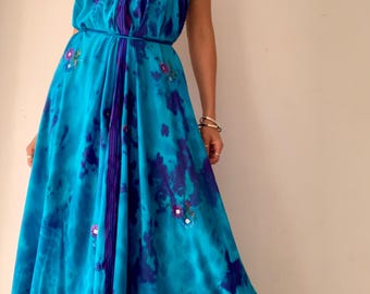May include: A floor-length halter dress in vibrant turquoise and deep blue tie-dye patterns. The garment features a deep V-neckline with dark purple trim, a thin waist tie, and small floral embroidery accents scattered across the fabric.