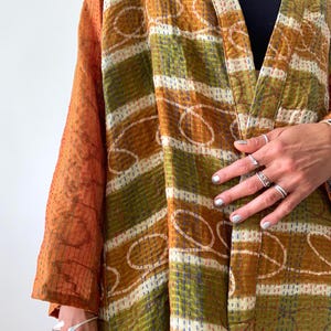 Op de afbeelding: Een patchwork kimono-jas in oranje, olijfgroen en wit. De jas heeft een gestreept patroon met gestikte details en een oranje mouw. Het openvallende kledingstuk is gemaakt van een zachte, getextureerde stof. Het ontwerp doet denken aan traditioneel patchwork.