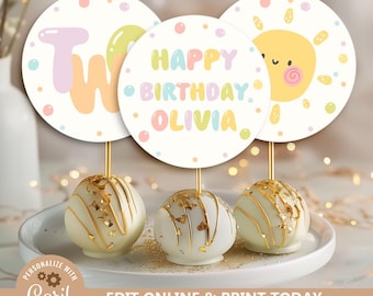 Puede incluir: Tres cake pops con detalles dorados y adornos decorativos. Los adornos son redondos con texto pastel que dice "Two", "Happy Birthday Olivia" y un sol sonriente. Los cake pops están en un plato blanco.