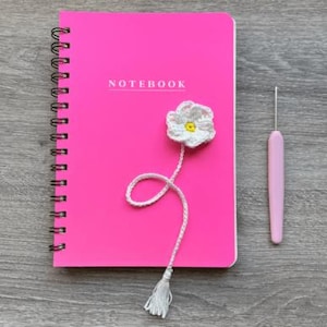 Puede incluir: Un cuaderno rosa brillante con espiral con la palabra "NOTEBOOK" impresa en la portada. Un marcapáginas de flores blancas de ganchillo con un centro amarillo y una borla está adjunto. También hay un ganchillo rosa.