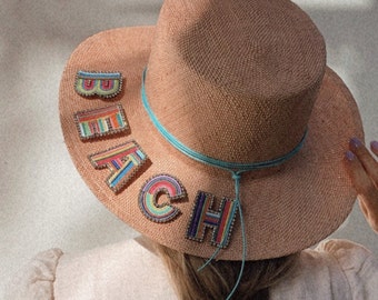 Pode incluir: Um chapéu de palha de aba larga com uma fita azul e uma coroa marrom. O chapéu é decorado com letras bordadas coloridas que soletram "BEACH".