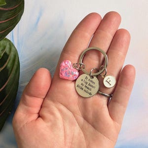 以下が含まれることがあります： ピンクのハートのチャームと「小さな心を教えるには大きな心が必要だ」というテキストが刻まれたシルバーのチャームが付いたシルバーのキーホルダーです。