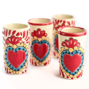 Puede incluir: Cuatro vasos de chupito de cerámica con base crema, cada uno adornado con un vibrante diseño de corazón rojo. Los corazones están rodeados de detalles azules y amarillos, y los bordes son rojos. Los vasos están sobre un fondo blanco.