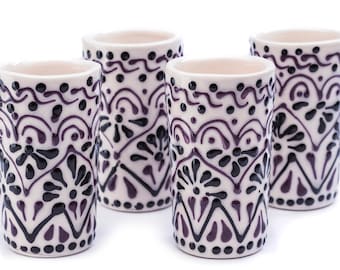 Puede incluir: Cuatro vasos de chupito de cerámica con base color crema y complejos diseños florales y geométricos en negro y morado. Los vasos son cilíndricos con un borde ligeramente acampanado.