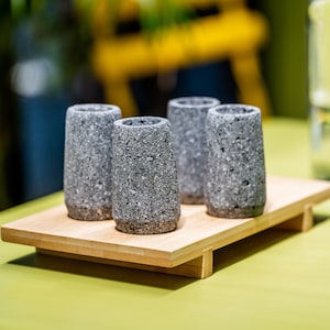 May include: Four gray stone shot glasses with a textured surface, arranged on a light brown bamboo tray. The shot glasses have a cylindrical shape with a slightly flared top. The tray is rectangular with a simple design.