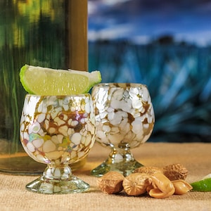May include: Two decorative shot glasses with a speckled pattern, topped with a lime wedge. A bottle of liquor is in the background, along with peanuts. The glasses have a unique design, perfect for serving tequila or other spirits.