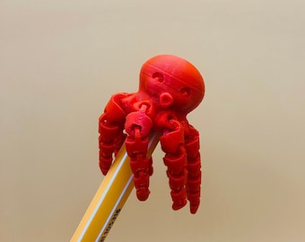 May include: A red 3D printed octopus toy perched on a yellow pencil. The octopus has eight tentacles and a round head with two small black eyes.