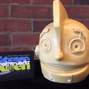 Puede incluir: Una cabeza de robot de color beige con una gran aleta puntiaguda en la parte superior. El robot tiene ojos grandes y redondos y una amplia sonrisa con dientes. La cabeza está hecha de un material liso, similar al plástico.