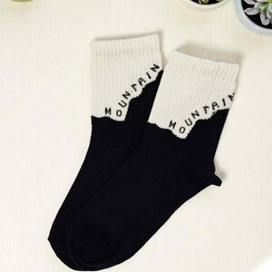 May include: A pair of black socks with white ribbed cuffs. The white cuffs feature the word "MOUNTAIN" in black block letters. The socks are displayed on a white surface, with small potted succulents in the background.