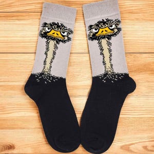 Könnte beinhalten: Ein Paar Socken mit einem Straußen-Design. Die Socken haben einen grauen oberen Teil, einen schwarzen Fuß und eine detaillierte Illustration eines Straußenkopfes mit gelbem Schnabel und wütenden Augen.