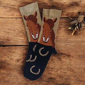 May include: A pair of socks with a horse theme. The socks feature a brown horse head on a tan background, with a navy blue cuff and horseshoe design. The socks are displayed on a wooden surface.