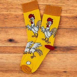 May include: A pair of yellow socks with a rooster design. The socks feature a red and white rooster on each sock. The socks are displayed on a wooden surface.