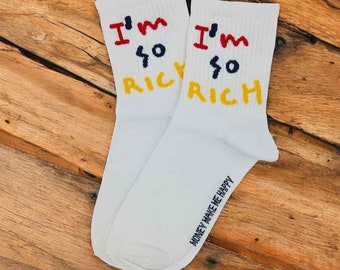 May include: A pair of white socks with the phrase "I'm so rich" written in red, blue, and yellow on each sock. The socks have the phrase "Money make me happy" written in blue on the bottom of one sock.