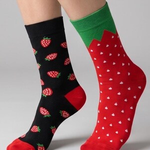 May include: A pair of crew socks featuring mismatched designs. The left sock is black with a repeating pattern of red strawberries and has a red heel and toe. The right sock is red with white speckles, a green cuff, and a green leaf-shaped trim.