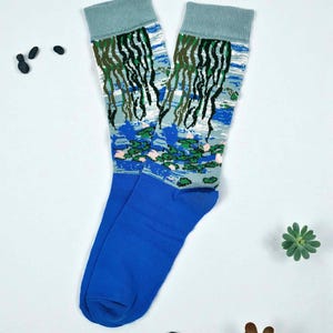 May include: A pair of blue socks with a water lily design. The socks have a blue cuff and toe, with a watercolor-style print of a pond scene featuring water lilies and weeping willow trees. The socks are laying on a white surface.